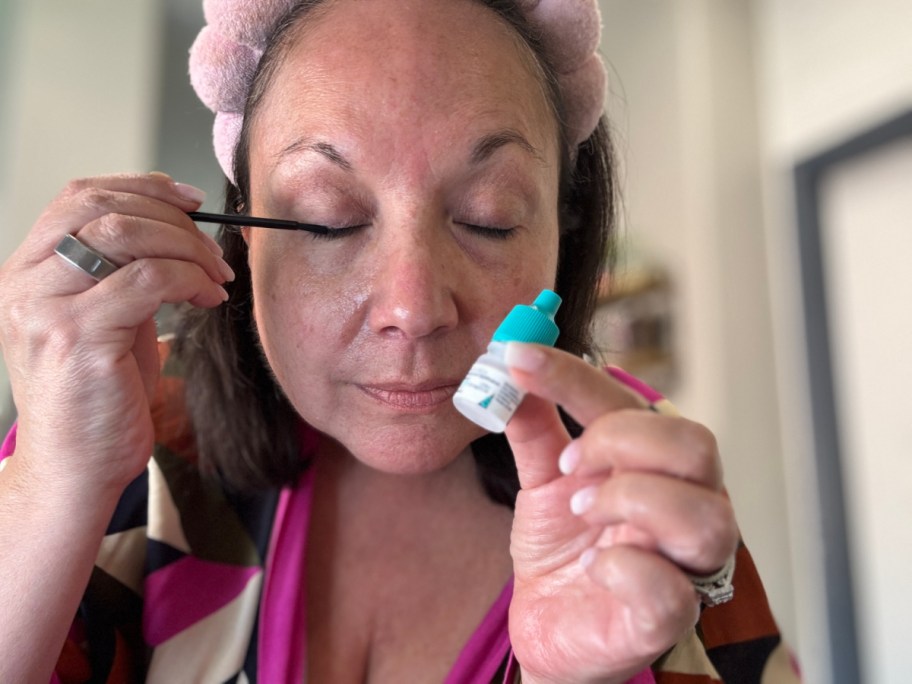 Woman applying eye lash serum with eyes closed, wearing a pink headband. Holds eye serum bottle.