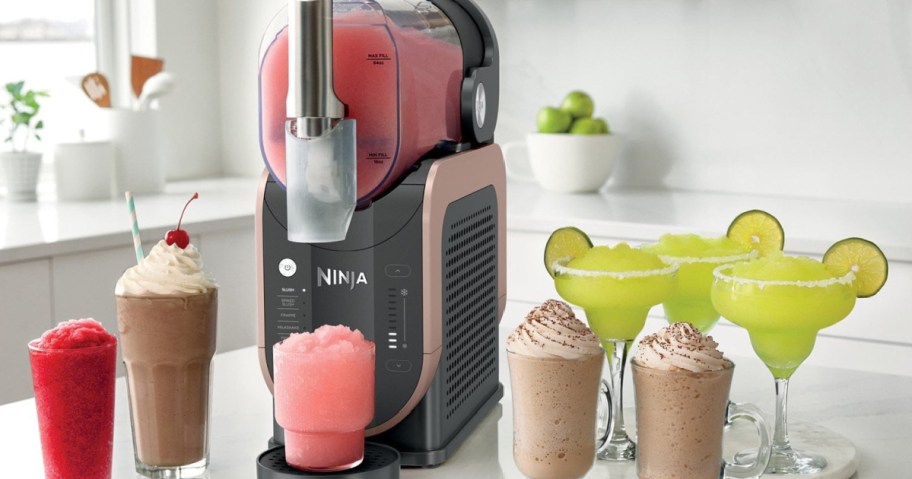 A sleek gray and pink drink machine on a white countertop dispenses a pink slush. Surrounding it are colorful drinks, including frappes and margaritas.