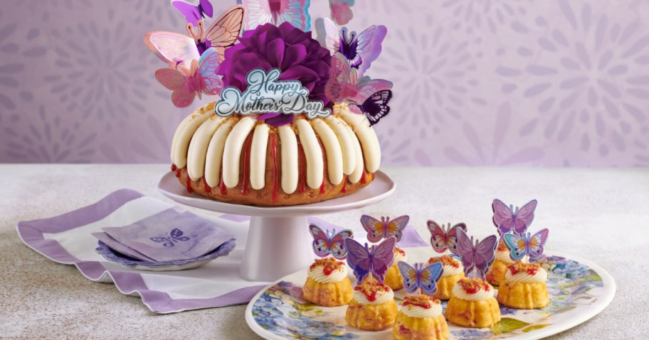 Mother's Day cake with white icing and a purple flower, topped with colorful butterfly decorations. Smaller cakes with butterfly toppers on a floral plate.