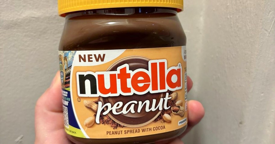 A hand holds a jar of Nutella Peanut, featuring a yellow lid and brown label. The label reads "NEW" and "peanut spread with cocoa," with images of peanuts.