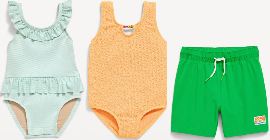 Children's swimwear set: pastel green ruffled swimsuit, orange textured one-piece, and vibrant green swim shorts with a white drawstring.