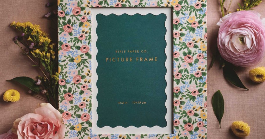 A floral patterned picture frame with a green center insert, surrounded by pink and yellow flowers on a tan surface.