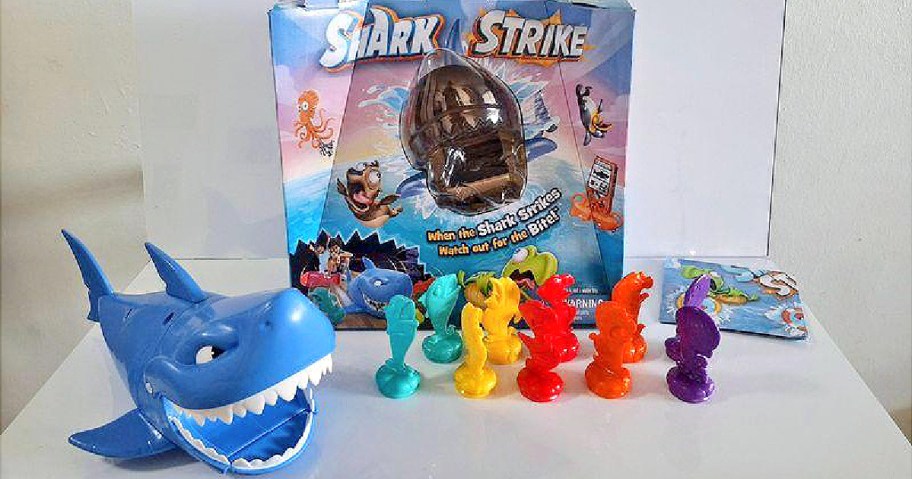 A colorful game set featuring a blue shark head, vibrant plastic figurines in various colors, and a box labeled "Shark Strike" with playful graphics.