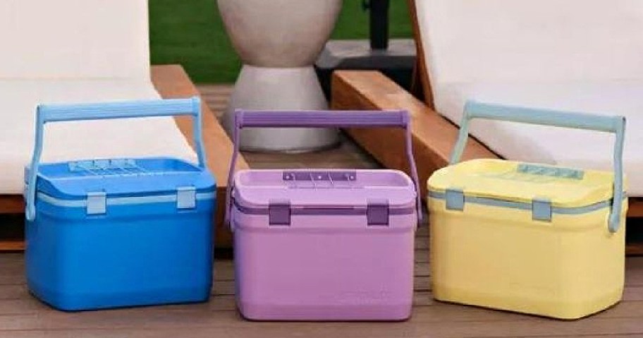 Three colorful cooler boxes in blue, purple, and yellow with sturdy handles are placed on a wooden patio.