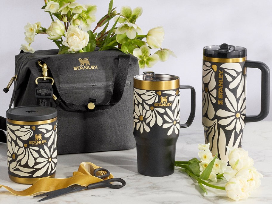 Stylish floral Stanley drinkware set with gold accents, featuring a mug, tumbler, and travel cup. A matching gray bag and white flowers complement the scene.