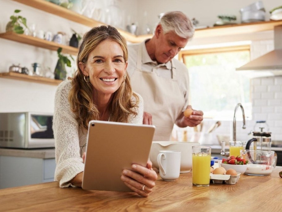 woman holding an iPad next to a man making food in a kitchen