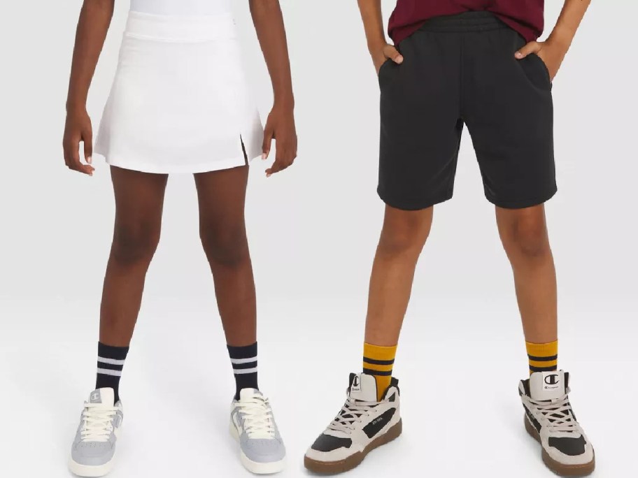 Two children stand side by side. One wears a white skirt, striped socks, and sneakers. The other wears black shorts, yellow socks, and high-top sneakers.