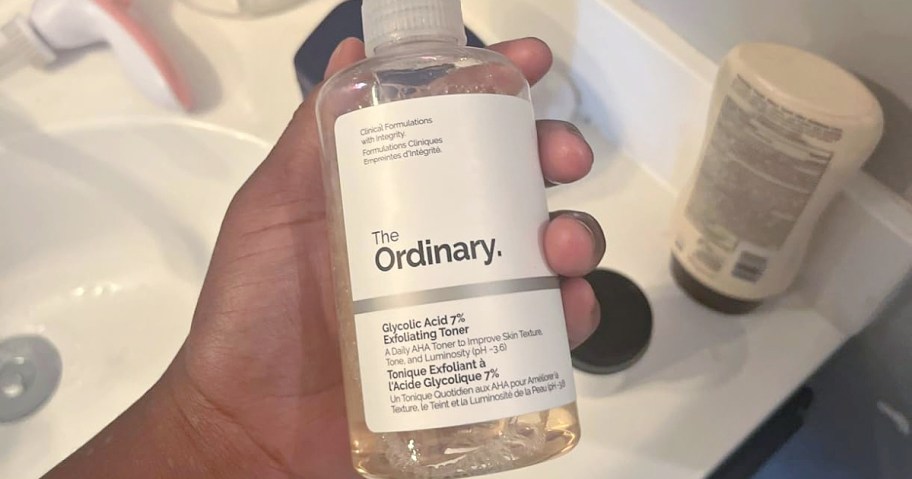 A hand holds a clear bottle of The Ordinary Glycolic Acid 7% Exfoliating Toner against a bathroom sink backdrop.
