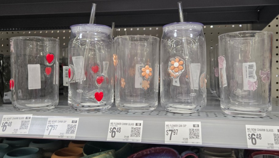 Clear glass tumblers on a store shelf, each decorated with colorful stickers of hearts, flowers, or bows. Prices range from $6.48 to $7.97.