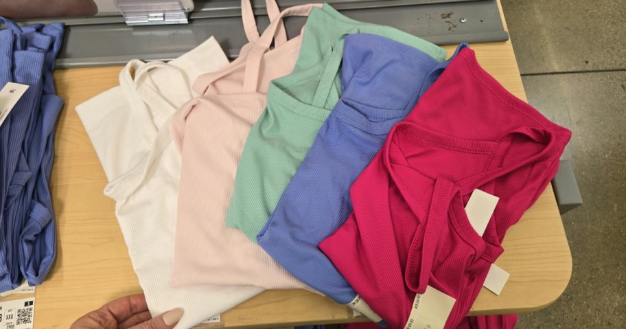 A variety of colorful tank tops are neatly stacked on a wooden store display. The tops are in white, pink, green, blue, and bright pink.