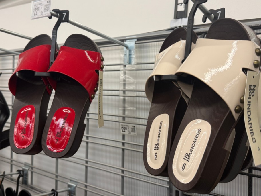 Hanging on a store display are two pairs of "No Boundaries" sandals, one red and one beige, both with glossy straps and wooden soles.