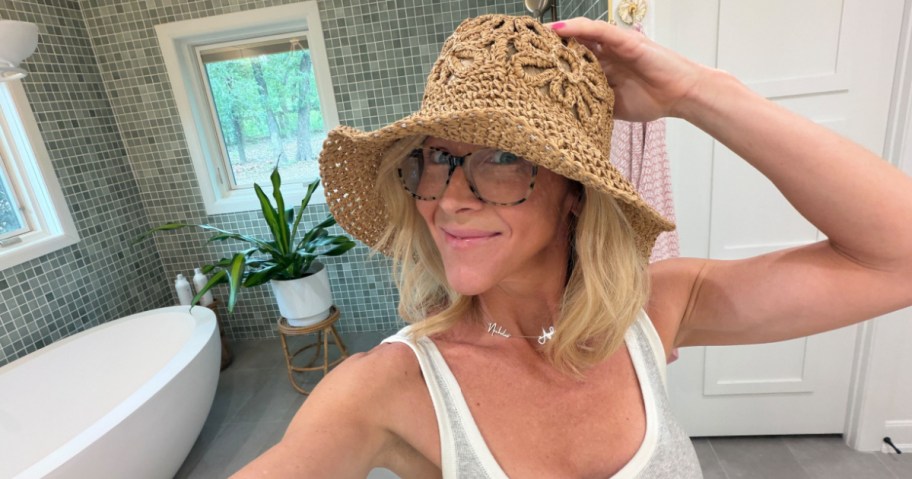 Woman in a bathroom wearing a beige woven sun hat and glasses, smiling at the camera. A white bathtub and green plant are in the background.
