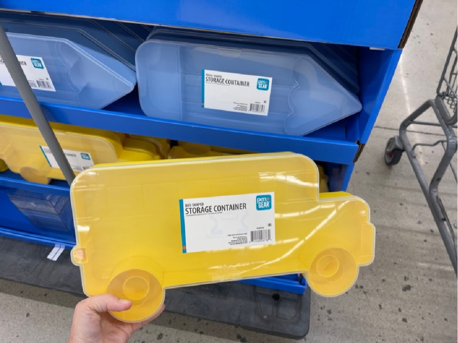 Yellow bus-shaped storage container held in a hand, displayed on a store shelf with similar blue containers. Labels read "Bus Shaped Storage Container."