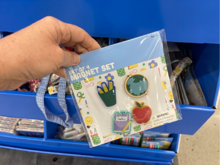 A hand holds a pack of four school-themed magnets, featuring a pencil cup, globe, notebook, and apple. Display bins with similar products are visible in the background.