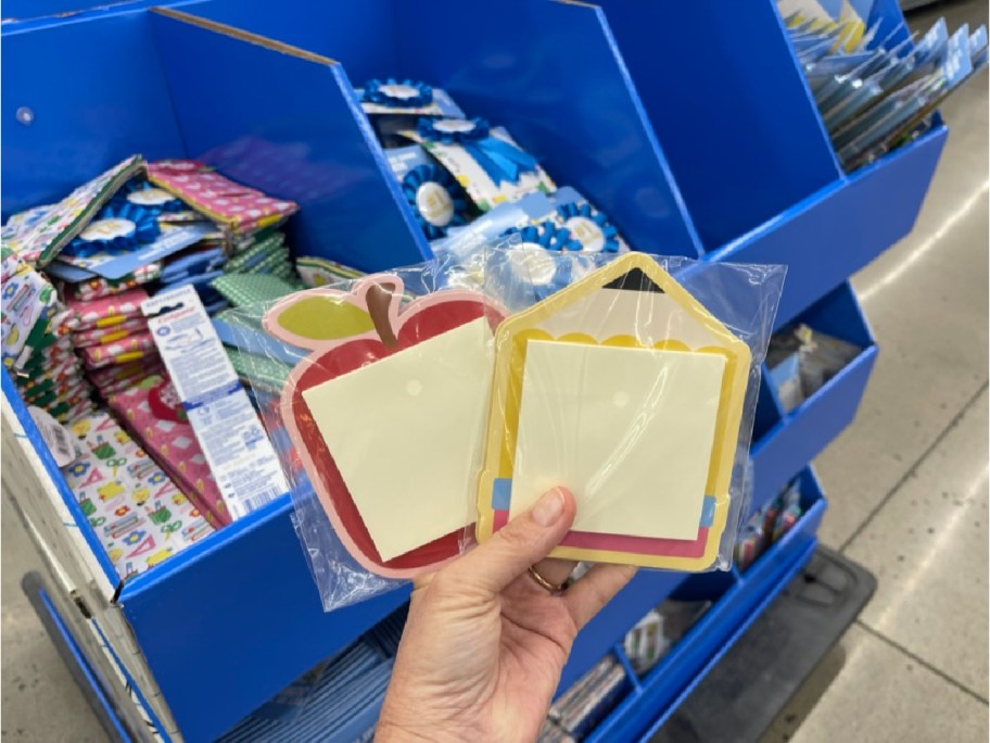 A close-up of a hand holding two packs of decorative sticky notes in an apple and house shape, in front of a display of assorted stationery in a store.