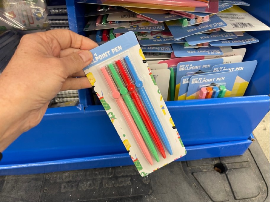 A hand holds a pack of four colorful ballpoint pens&mdash;red, green, pink, and blue. They are displayed against a blue basket full of similar pen packs.