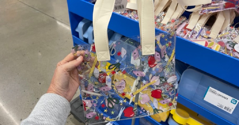 A hand holds a small, transparent tote bag with white handles, featuring colorful patterns of stars, lips, scissors, and flowers. The background shows store shelves filled with similar bags and other containers.
