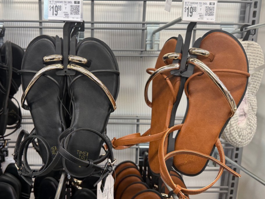A pair of sandals displayed on a rack; one pair is black with metallic accents, and the other pair is brown with similar design elements.