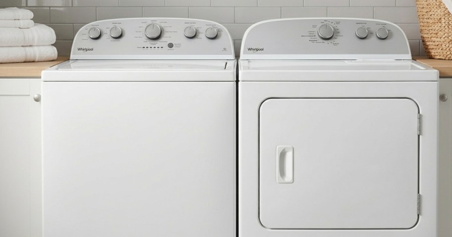A white top-loading washing machine and dryer sit side by side in a laundry room. The countertop above holds folded towels and a wicker basket.