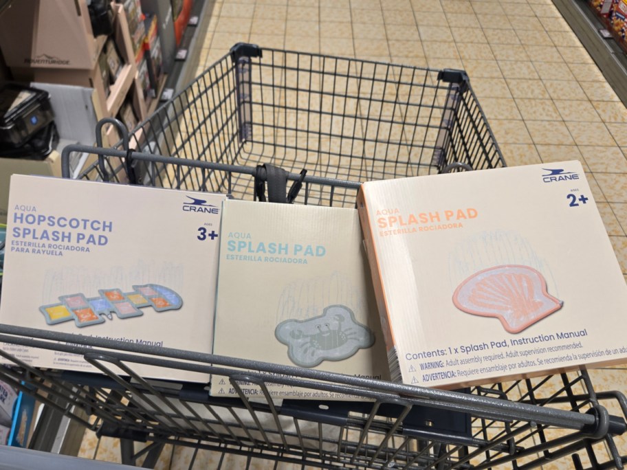 Shopping cart filled with three boxed Aqua splash pads by Crane. Each box shows different designs suitable for ages 2+. Store aisle in the background.