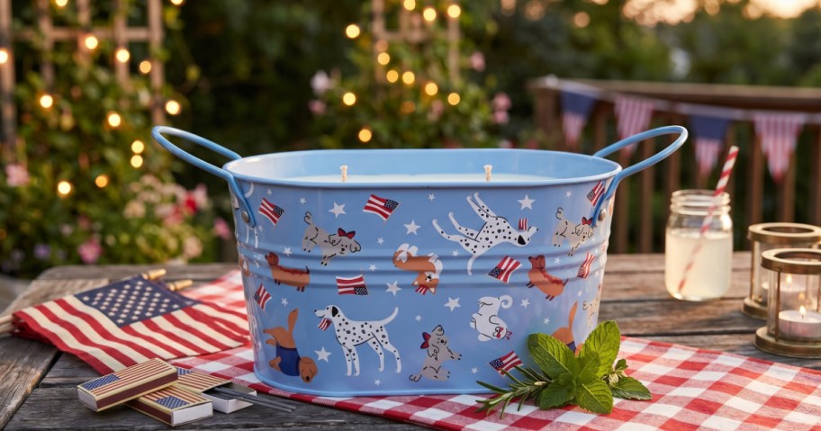 A festive metal citronella candle with patriotic dog illustrations sits on a picnic table, surrounded by American flags, string lights, and a jar of lemonade at dusk.