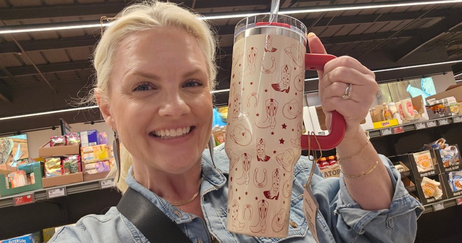 A smiling person in a denim shirt holds a large, decorated tumbler with a red handle in a grocery store aisle.