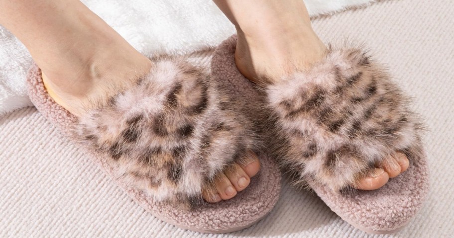 Feet wearing cozy, light pink slippers with fuzzy, fur-like leopard print on a textured cream carpet.