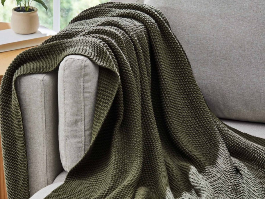 A dark green chunky knit throw blanket draped over the arm of a light gray sofa.
