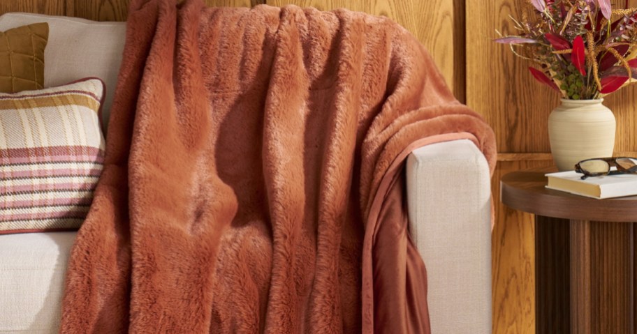 A plush, rust-colored faux fur throw blanket draped over a light beige sofa, positioned next to throw pillows and a small wooden side table with a book and flowers.
