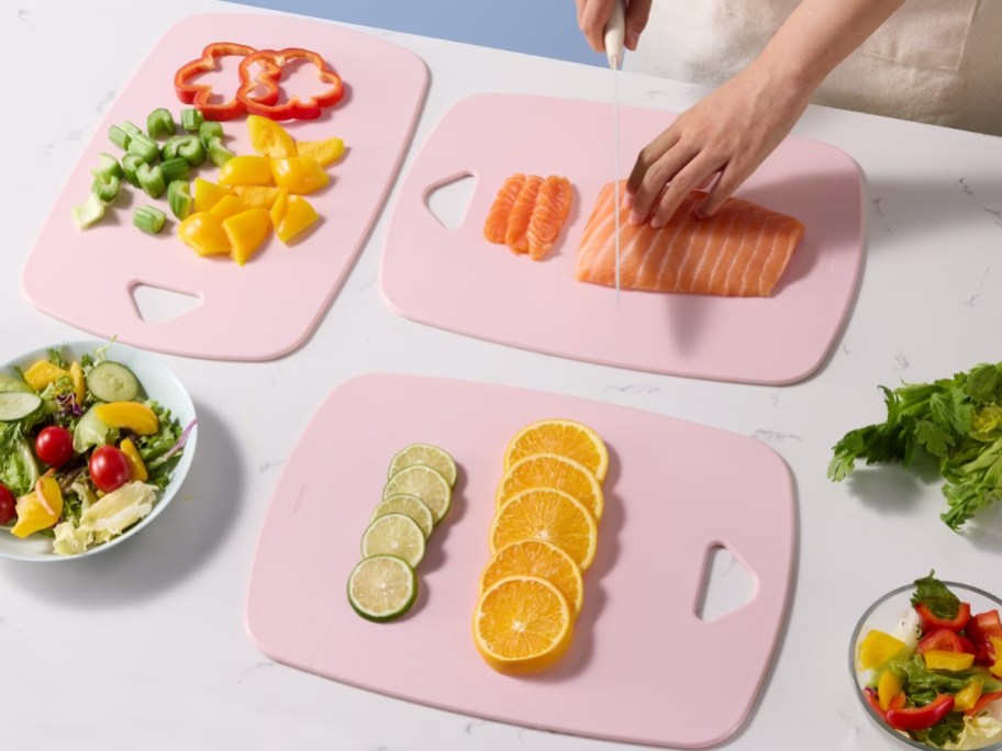 three pink plastic cutting boards on kitchen counter with vegetables, meat, and fruit on boards