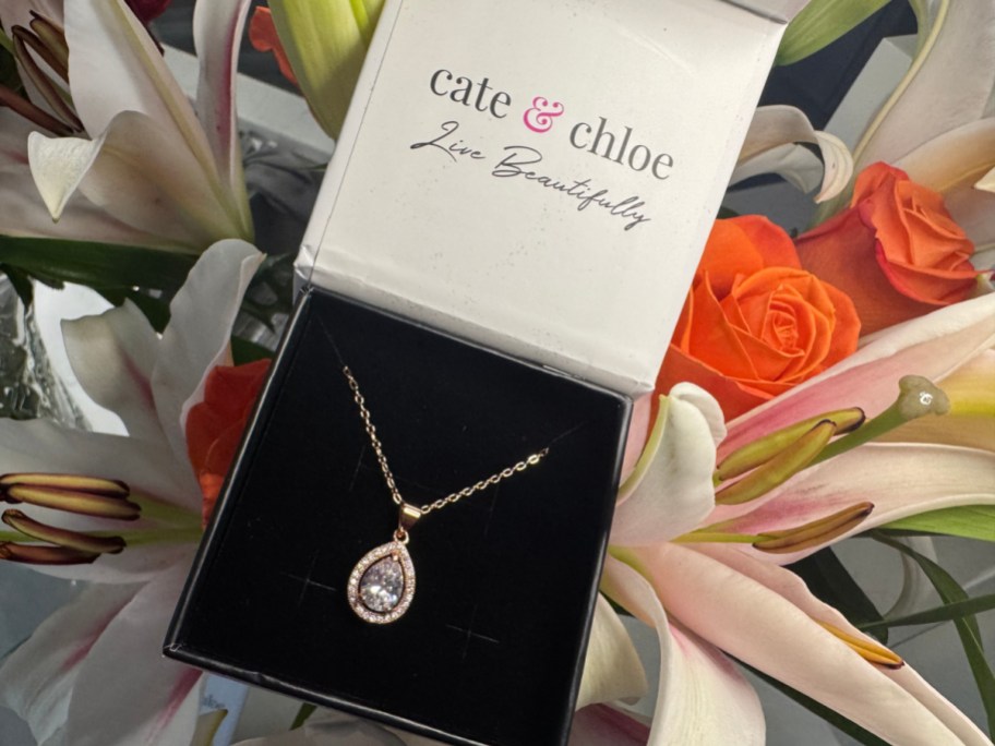 A gold-plated teardrop halo pendant necklace resting inside an open Cate & Chloe gift box surrounded by flowers.