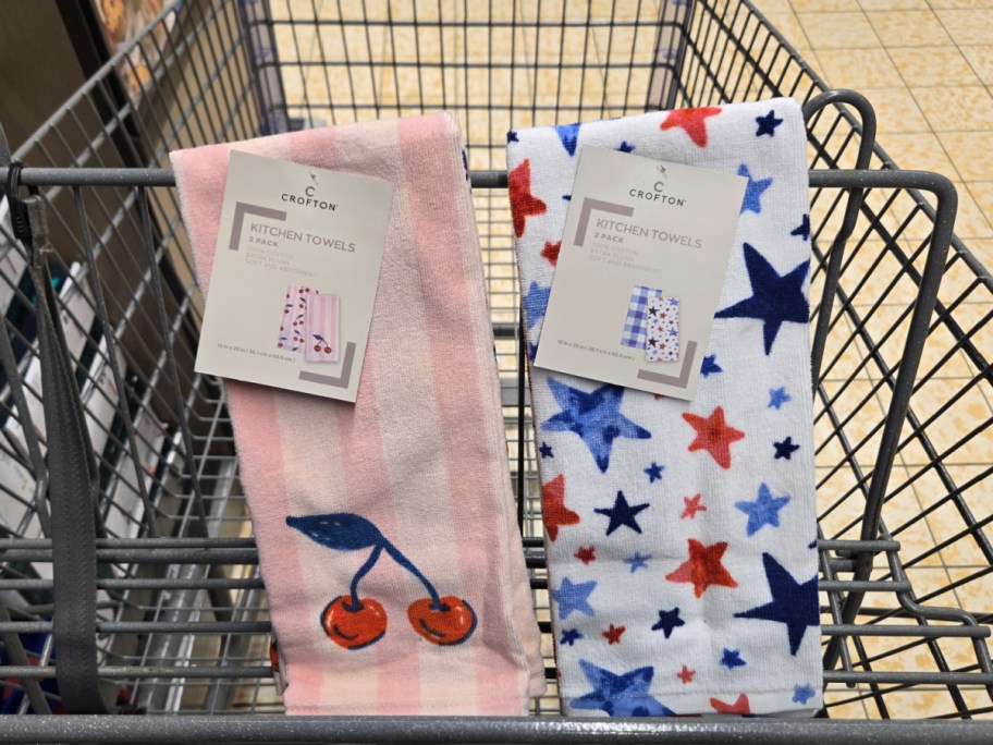 Two packs of Crofton kitchen towels lie in a shopping cart. One is pink with cherries, the other white with red and blue stars.