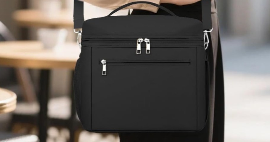 A person holding a black insulated lunch bag with multiple zippered compartments, standing near outdoor furniture.