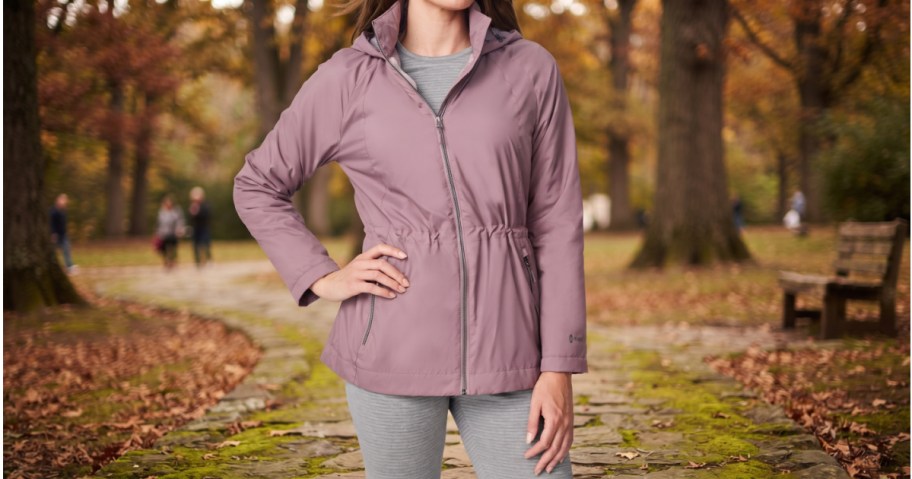 A person wearing a mauve anorak jacket and gray pants while standing on a stone path in an autumn park.