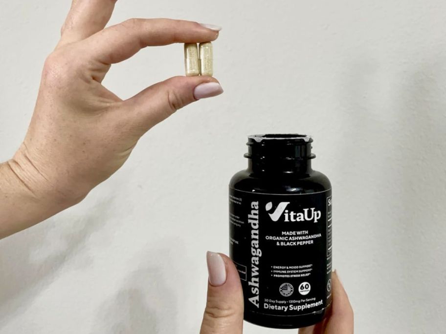 Hand holding two VitaUp Ashwaganda capsules over a bottle