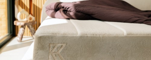 A cozy, sunlit bedroom with a beige mattress featuring a "K" logo on the side. A dark brown blanket is draped on top, and a rustic wooden stool is beside it.