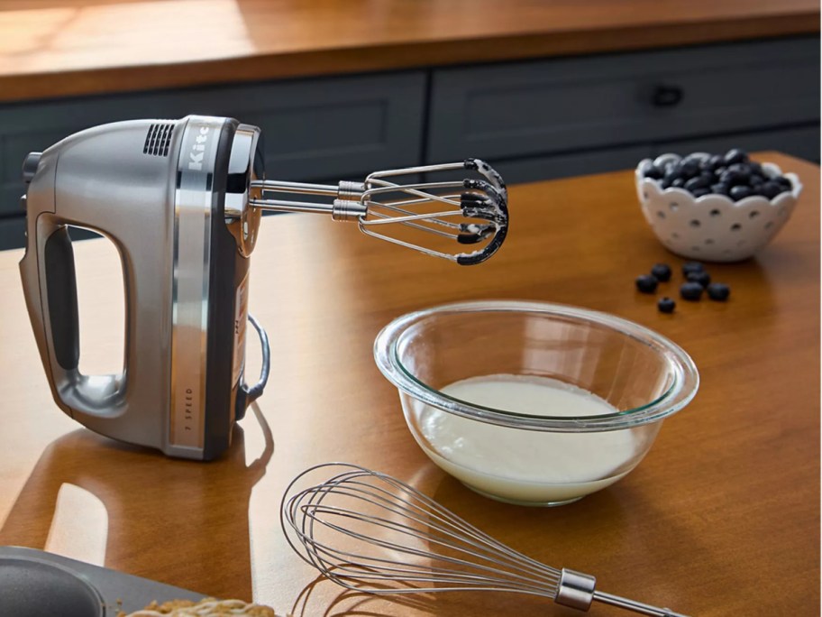 A silver KitchenAid hand mixer resting on a wooden countertop next to a glass mixing bowl, a wire whisk, and a bowl of blueberries.