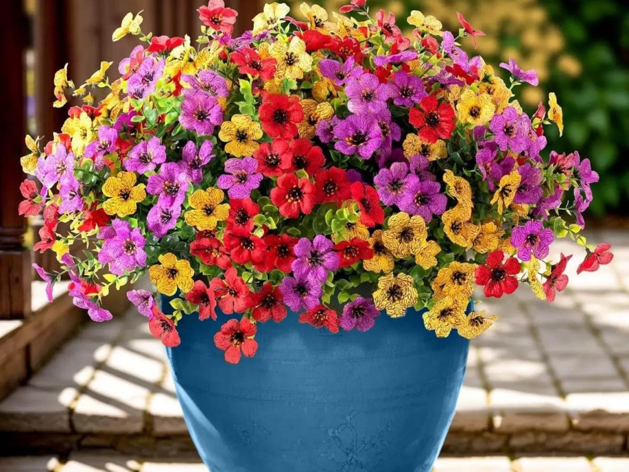 A vibrant bouquet of multicolored flowers, including red, pink, purple, and yellow blooms, arranged in a blue pot. The background features a sunny outdoor setting with greenery.