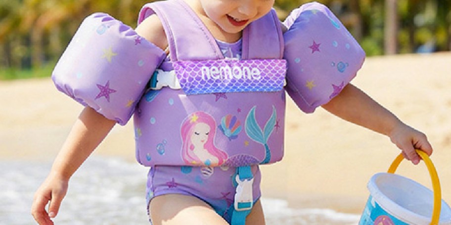 A child wearing a purple flotation device with a mermaid design, walking on the beach and holding a bucket.