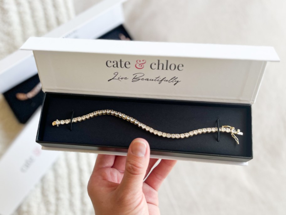 A hand holding an open white Cate & Chloe gift box containing a sparkling tennis bracelet.