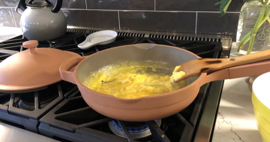 A pink our place always pan on a stove with scrambled eggs in the pan