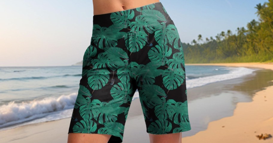 A person standing on a beach wearing black knee-length swim shorts with a green tropical leaf print.