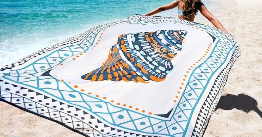 a woman walking on a beach with a large beach towel flowing behind her