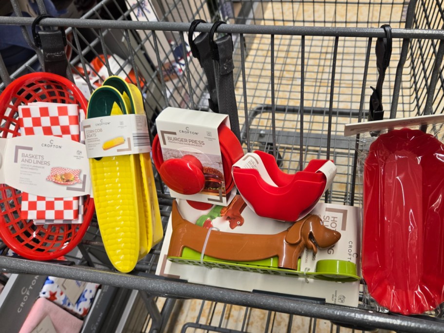 Shopping cart with colorful kitchen items: red food baskets, corn-shaped trays, a burger press, dachshund-shaped cutter, and a red baking dish.