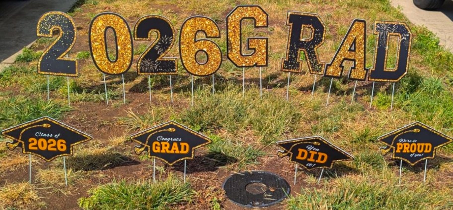 Lawn display with large glittery letters reading "2026 GRAD" and smaller signs shaped like graduation caps with messages like "Congrats GRAD" on grassy patch.