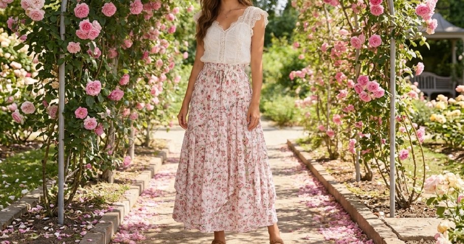 A woman standing in a flower garden, wearing a white lace top and a floral maxi skirt, surrounded by pink roses.