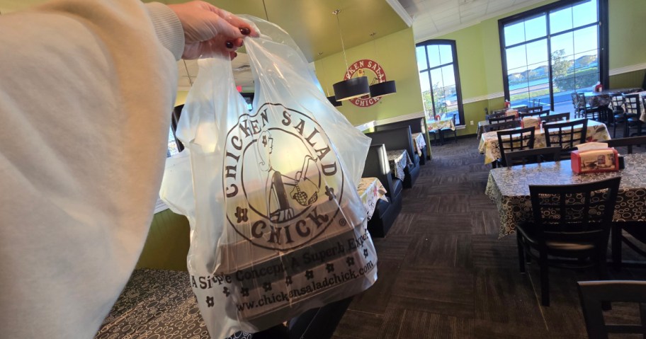 A person holds a Chicken Salad Chick takeout bag in a brightly lit restaurant with tables and chairs.