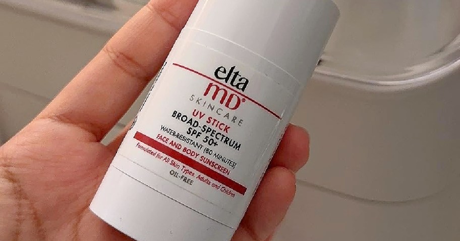 A hand holds a white tube of EltaMD UV Stick, labeled as broad spectrum SPF 50 sunscreen.