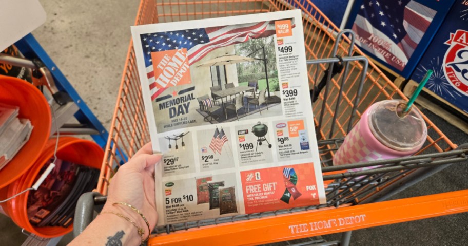 A person holds a Home Depot Memorial Day ad inside a shopping cart, featuring outdoor furniture and decorations.