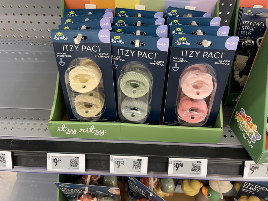 Three packages of Itzy Ritzy Itzy Paci silicone pacifiers displayed in a store bin, featuring cream, green, and pink colors.